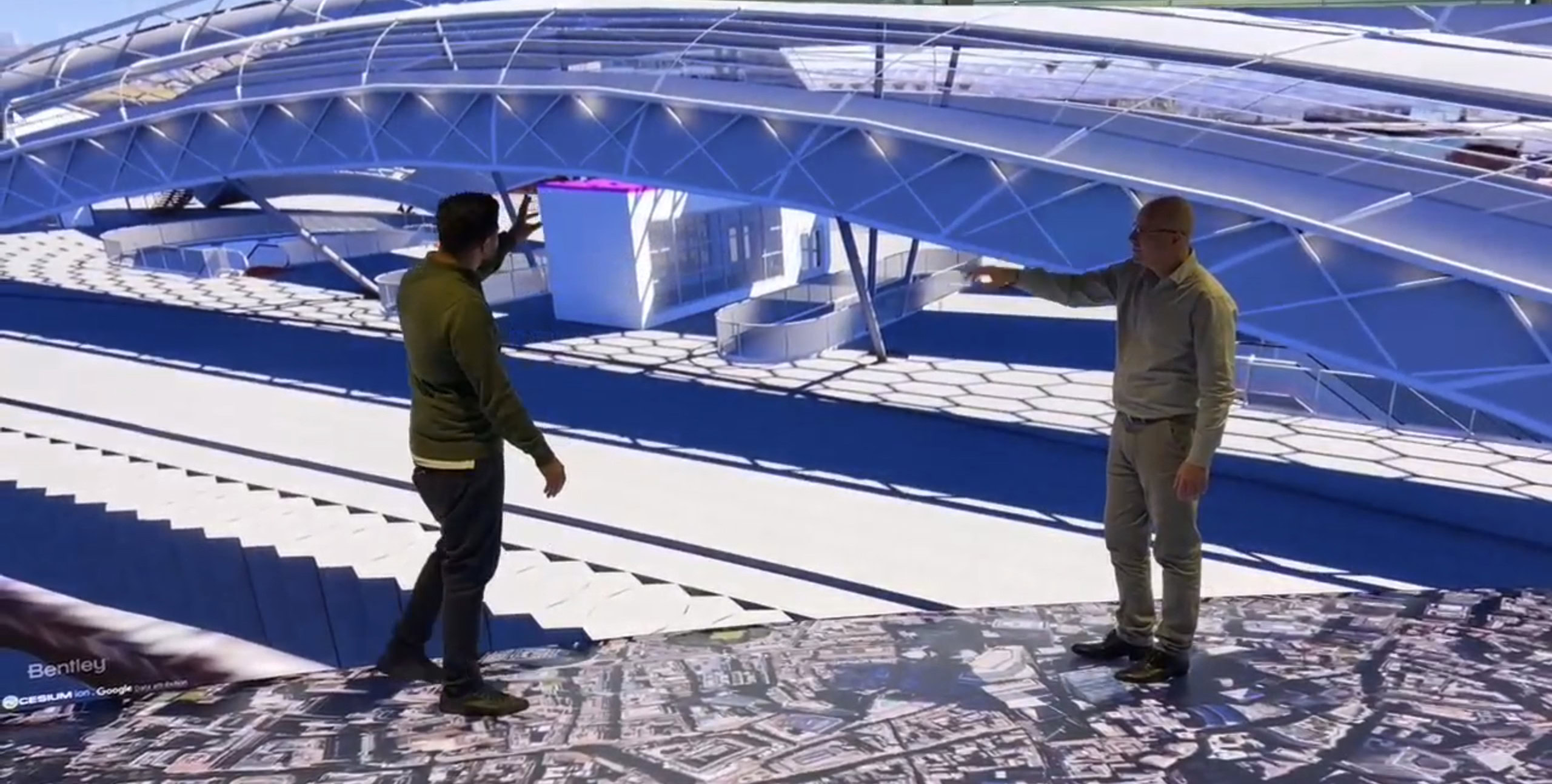 Two men standing on a digital map, powered by Bentley Systems' 3D Geospatial technology, view a large curved architectural structure on the screen, discussing its design elements.