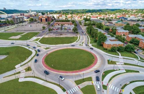 Aerial view of a traffic roundabout seamlessly integrated into urban road design, surrounded by cars, green spaces, and buildings in the background. | Bentley Systems | Infrastructure Engineering Software Company Aerial view of a traffic roundabout seamlessly integrated into urban road design, surrounded by cars, green spaces, and buildings in the background.